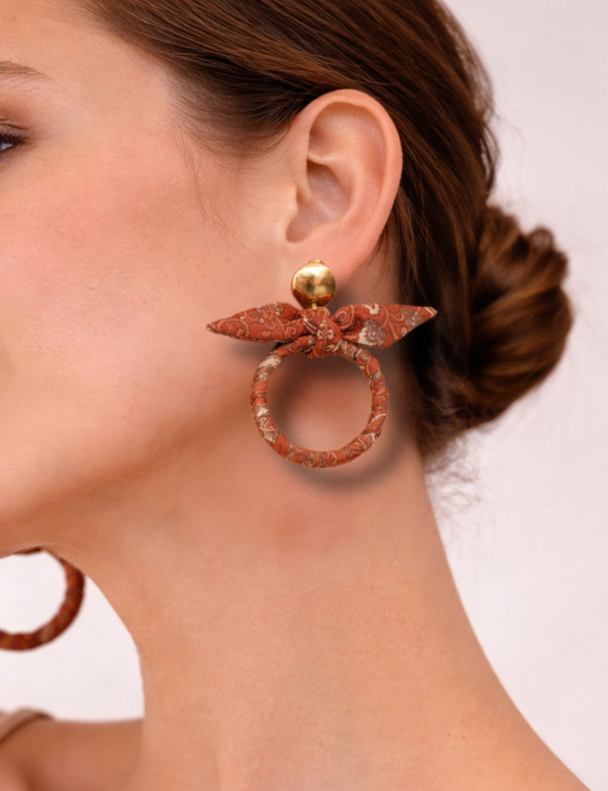 Close-up side view of a woman wearing orange tropical bow earrings with gold round studs and fabric-wrapped hoops