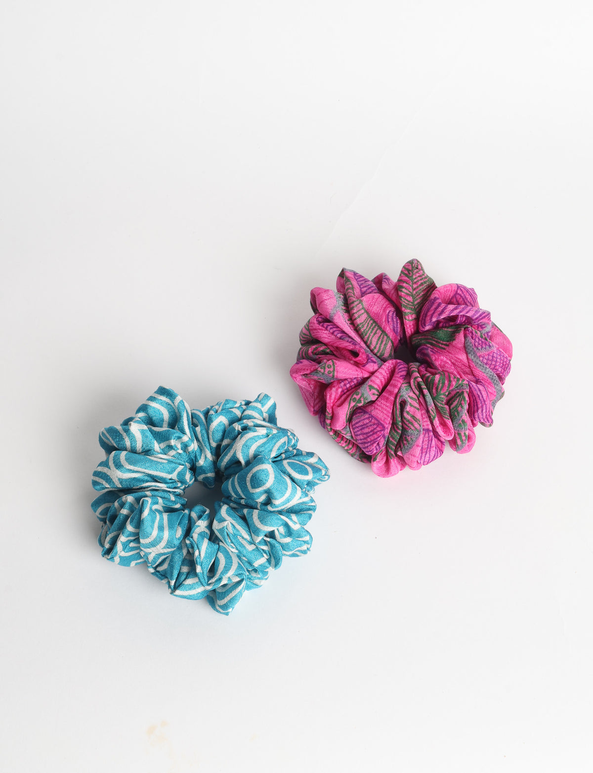 Set of two fabric scrunchies, one turquoise with beige abstract spots, one red with white linear pattern, flat lay on white