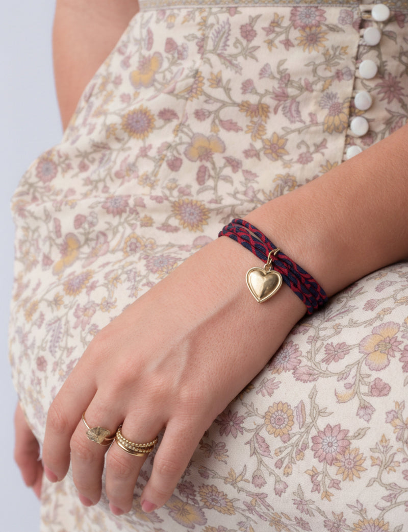 Red and navy twisted cord bracelet with gold heart charm worn on wrist with floral patterned dress and gold rings