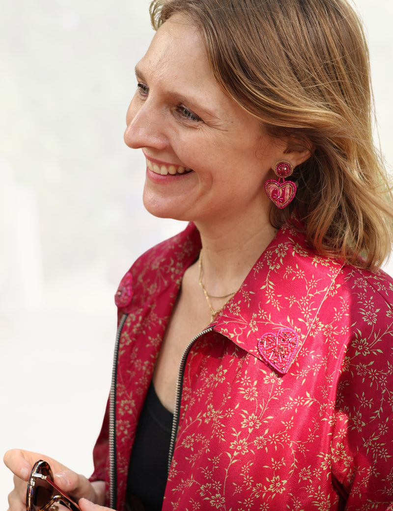Woman wearing pink abstract heart drop earrings and a red floral patterned jacket with a heart-shaped brooch, holding sunglasses