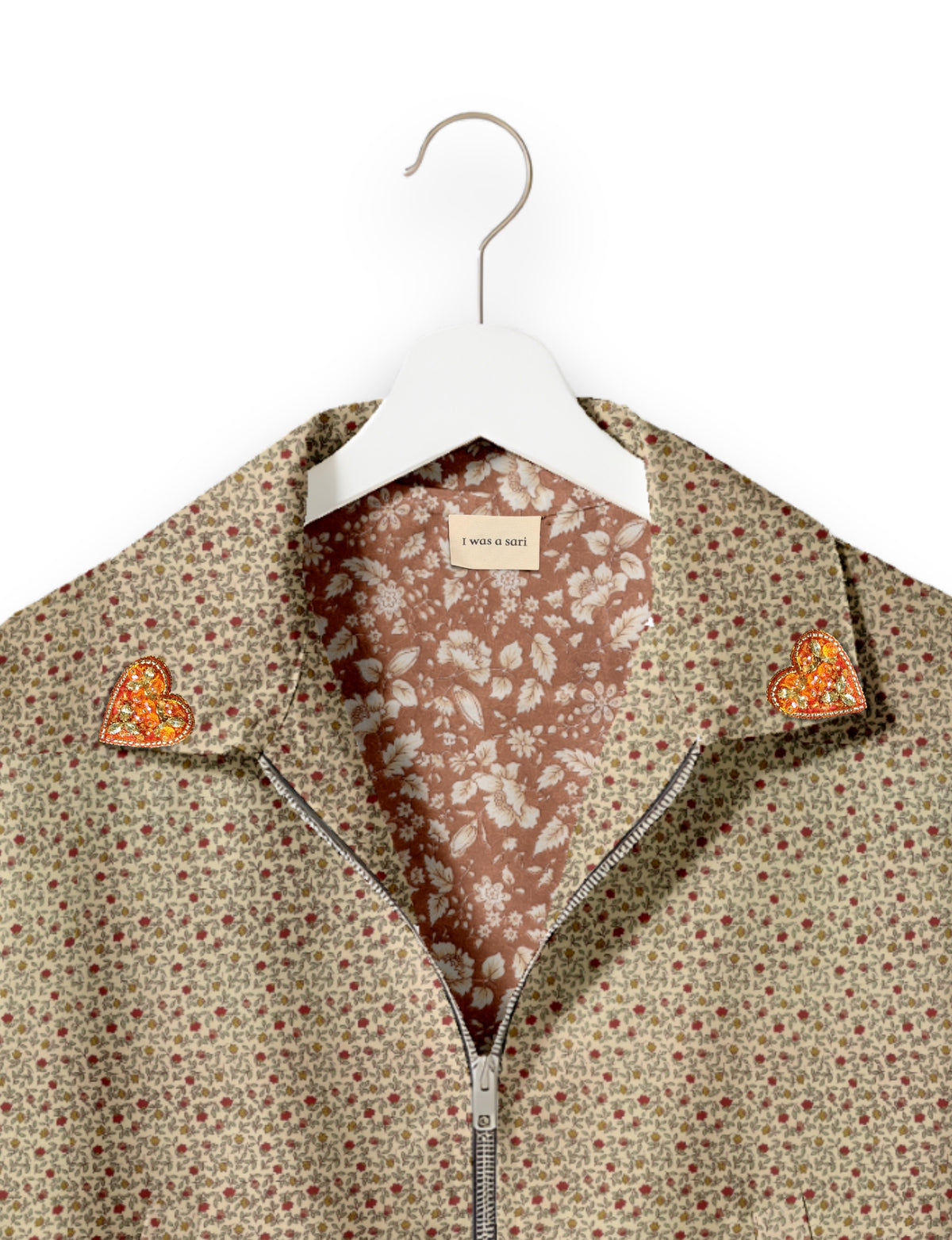 Beige floral bomber jacket with orange jeweled heart patches on collar, front zipper, and floral inner lining on white hanger