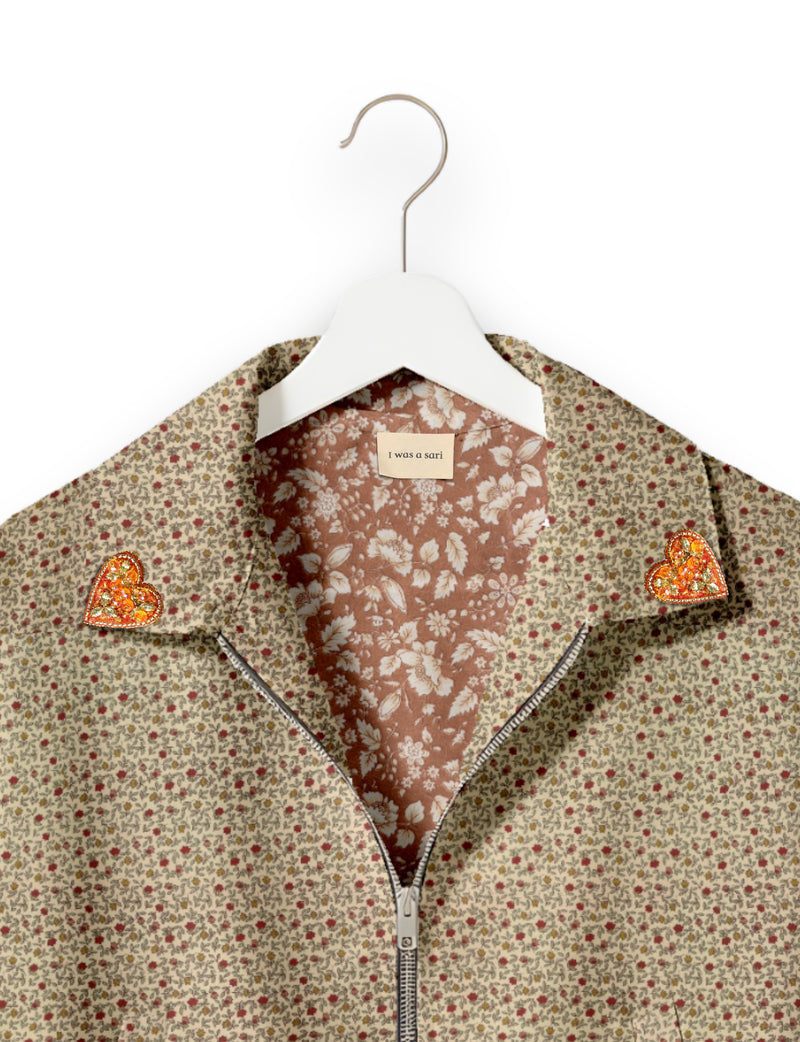 Beige floral bomber jacket with orange jeweled heart patches on collar, front zipper, and floral inner lining on white hanger