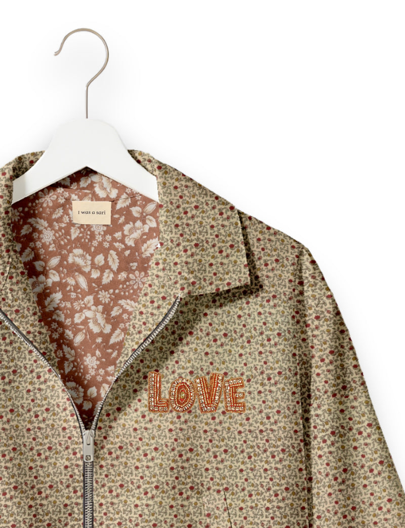 Beige floral bomber jacket with zipper front, embroidered "LOVE" patch, and contrasting floral inner lining on white hanger