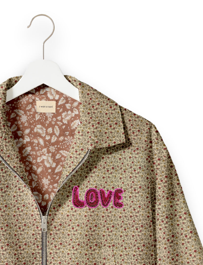 Beige floral bomber jacket with pink beaded "LOVE" patch on chest, floral inner lining, and silver zipper, hanging on white hanger