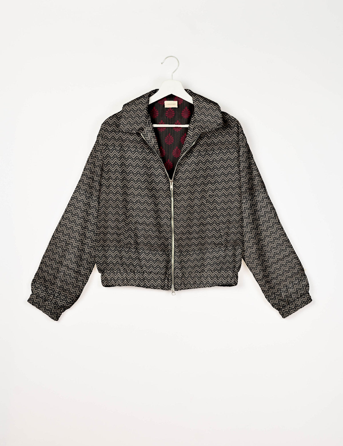 Beige bomber jacket with black geometric zigzag pattern, front zipper, elastic cuffs and hem, shown on hanger