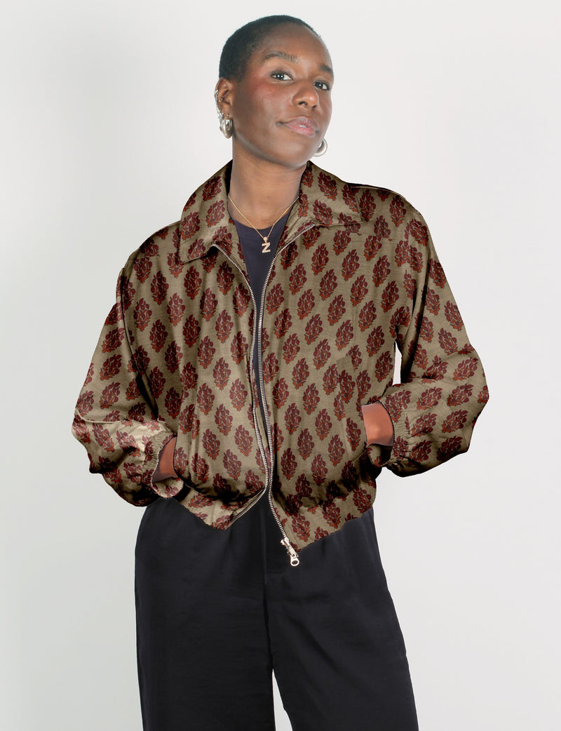 Woman wearing a beige bomber jacket with red tropical leaf pattern, front zipper, and elastic cuffs, paired with black pants