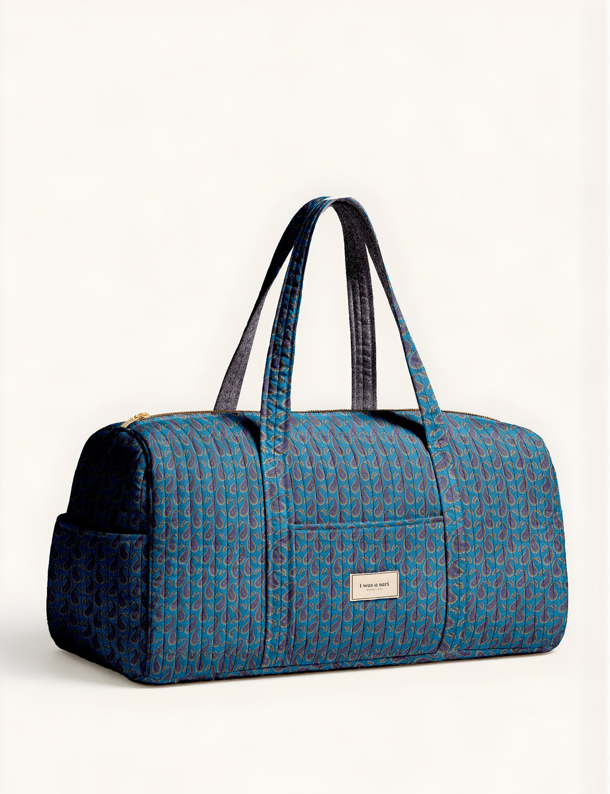 Woman holding a brown paisley quilted barrel bag with front zipper pocket, wearing light blue jeans and white sneakers
