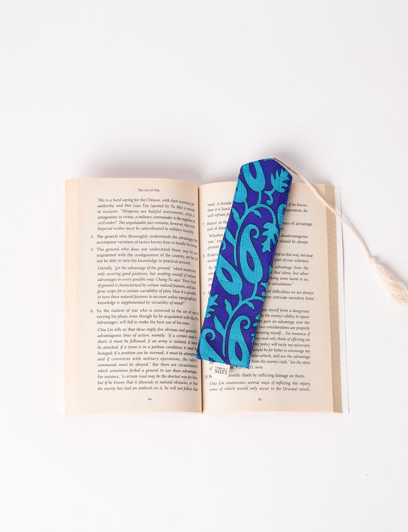 Blue fabric bookmark with turquoise floral pattern and beige tassel placed inside an open book flat on white background