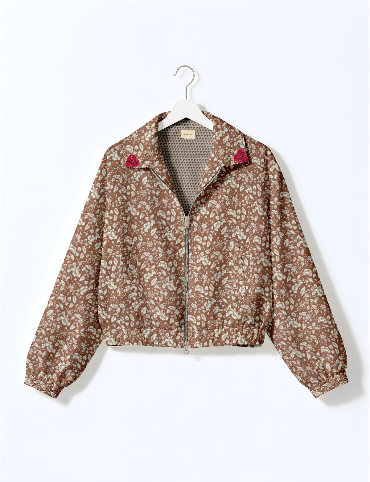 Brown floral bomber jacket with front zipper, elastic cuffs and hem, and pink heart patches on collar tips hanging on white hanger