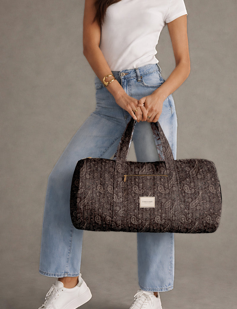 Brown paisley quilted bowling bag with front zipper pocket, held by person wearing white shirt and light blue jeans