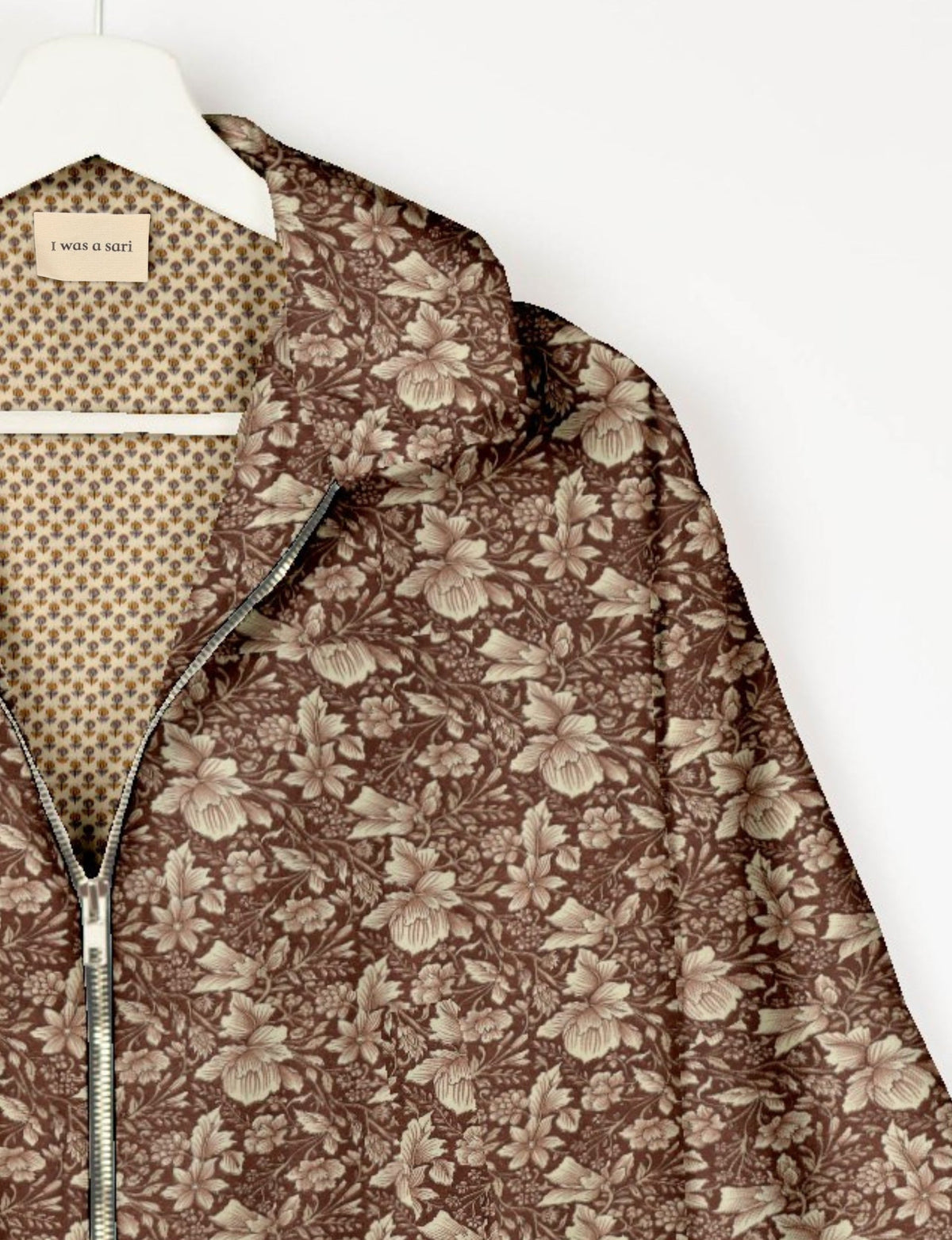 Brown floral patterned bomber jacket with silver zipper, hanging on white hanger, label reads "I was a sari" inside collar
