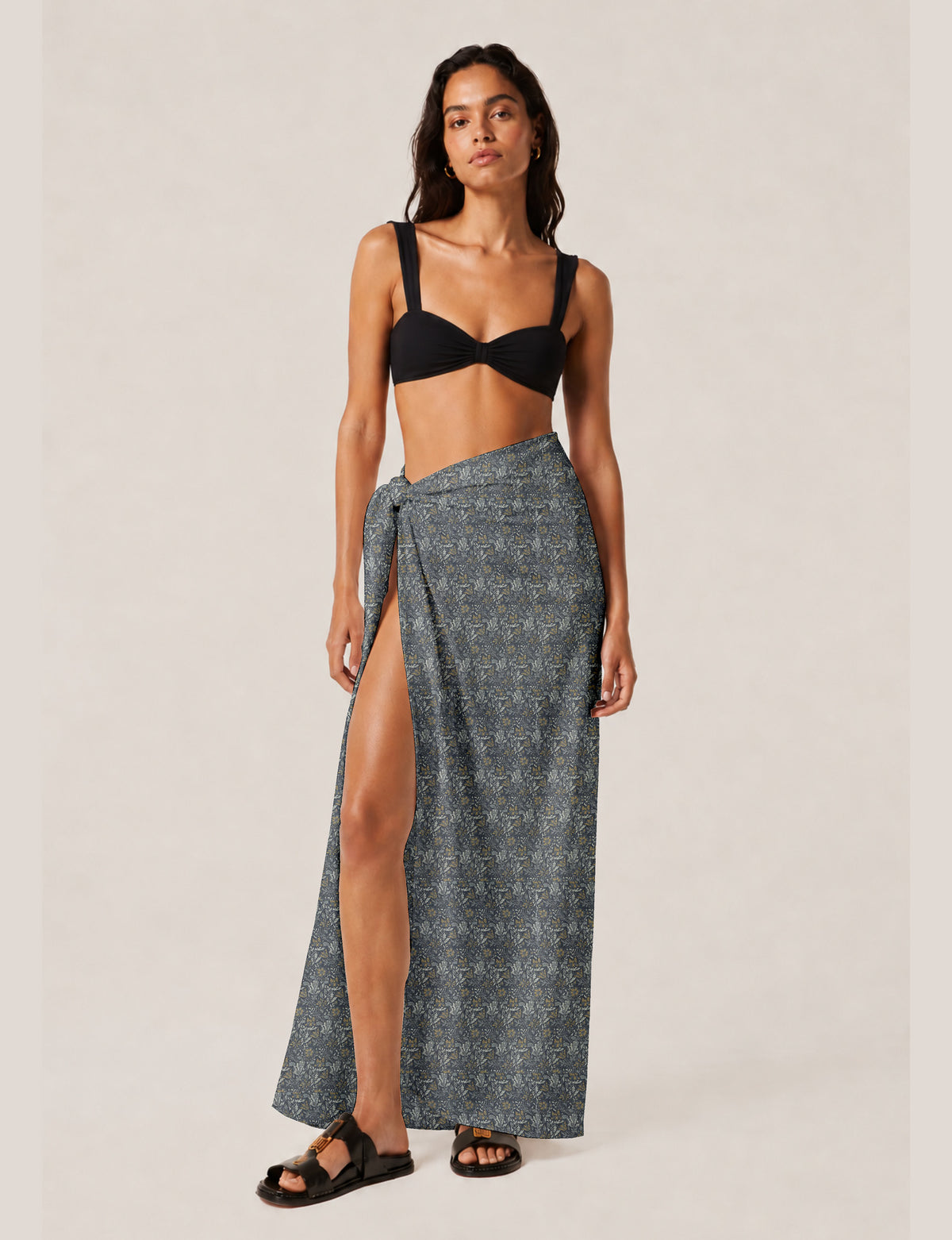 Woman wearing a grey tropical print pareo wrap skirt with a high slit, paired with a black bikini top and black sandals
