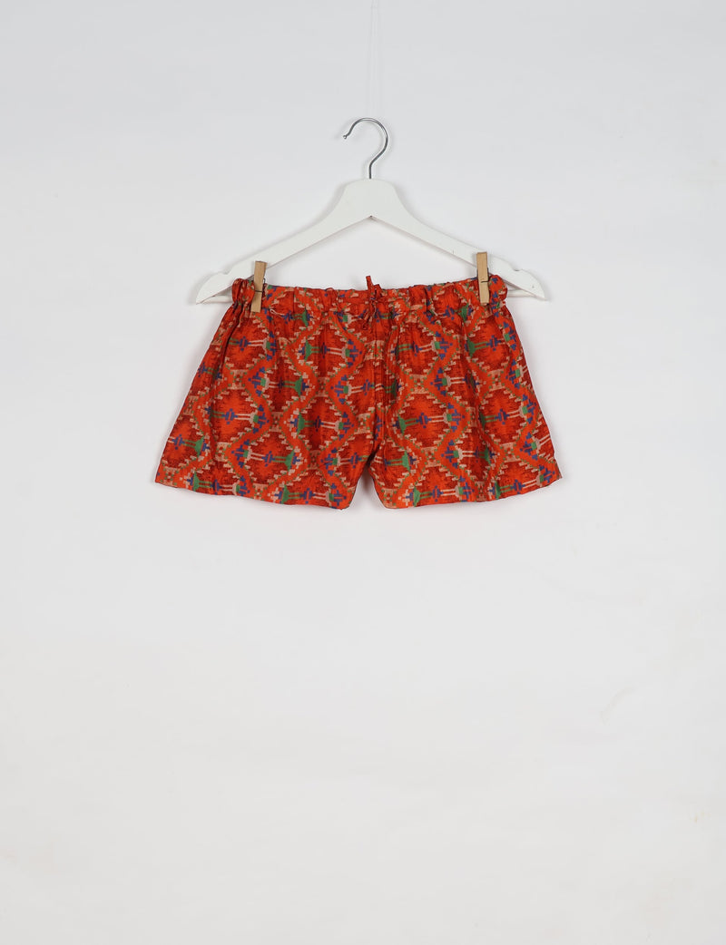 Orange ethnic patterned shorts with elastic waistband and drawstring, hanging on white hanger against white background