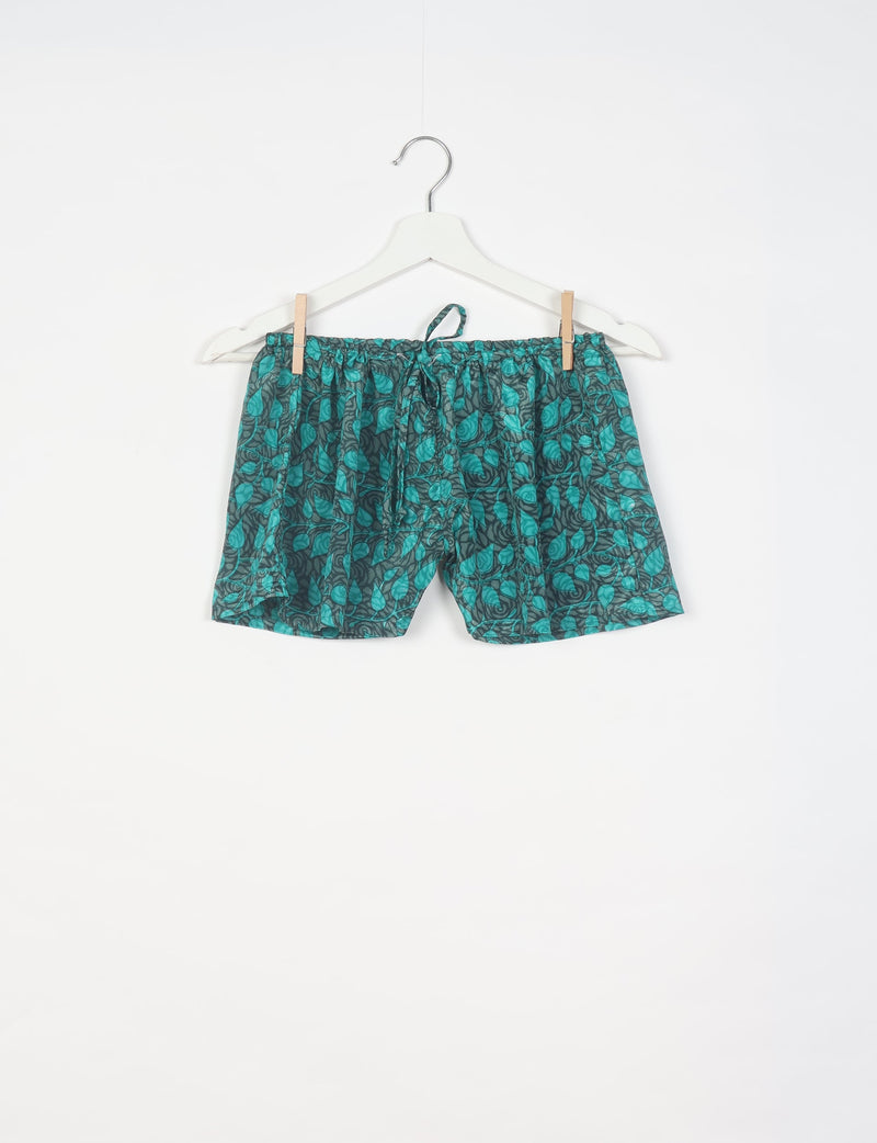 Green and black leaf-patterned shorts with elastic waistband and drawstring, hanging on white hanger against white background
