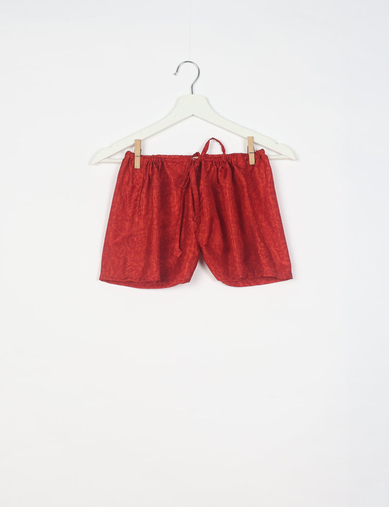 Red shorts with abstract pattern, elastic waistband, and drawstring, hanging on a white hanger against a white background