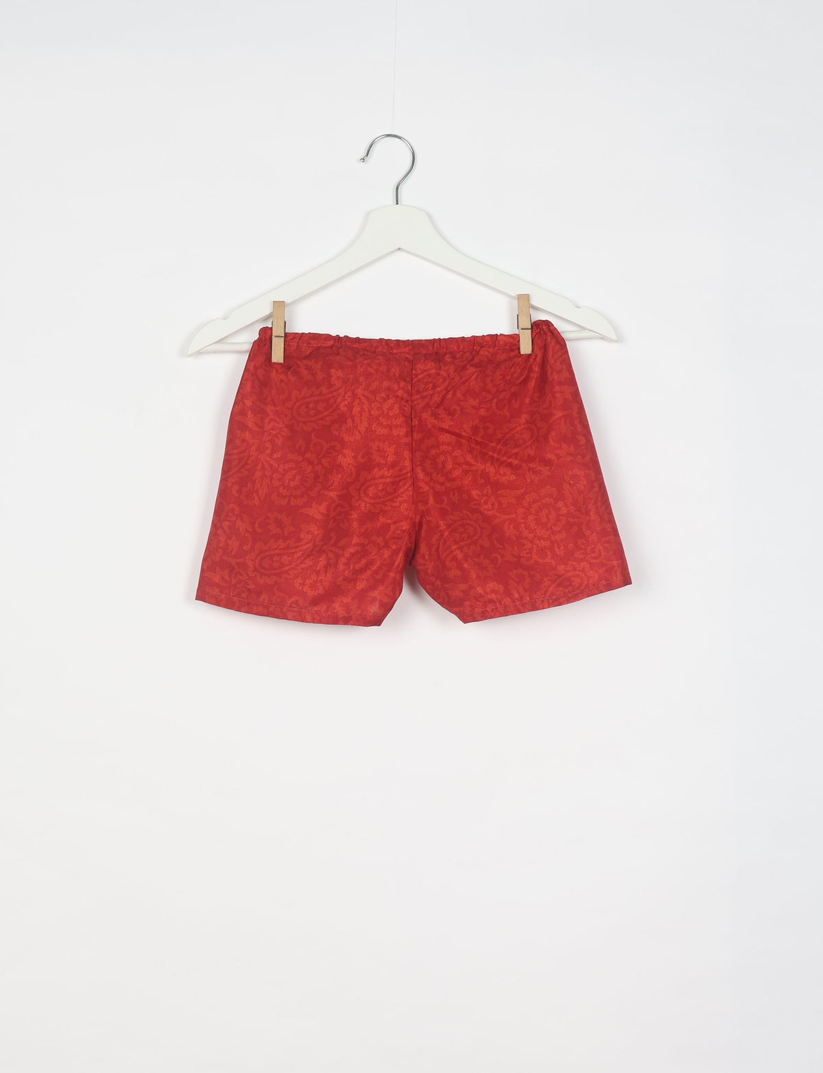 Red shorts with an abstract floral pattern, elastic waistband, hanging on a white hanger against a white background