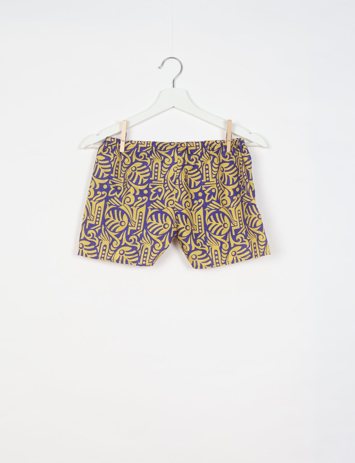 Purple shorts with yellow tropical abstract pattern hanging on white hanger against white background