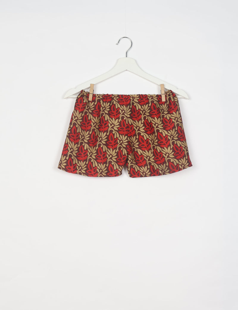 Red and beige leaf patterned shorts with elastic waistband hanging on white hanger against white background