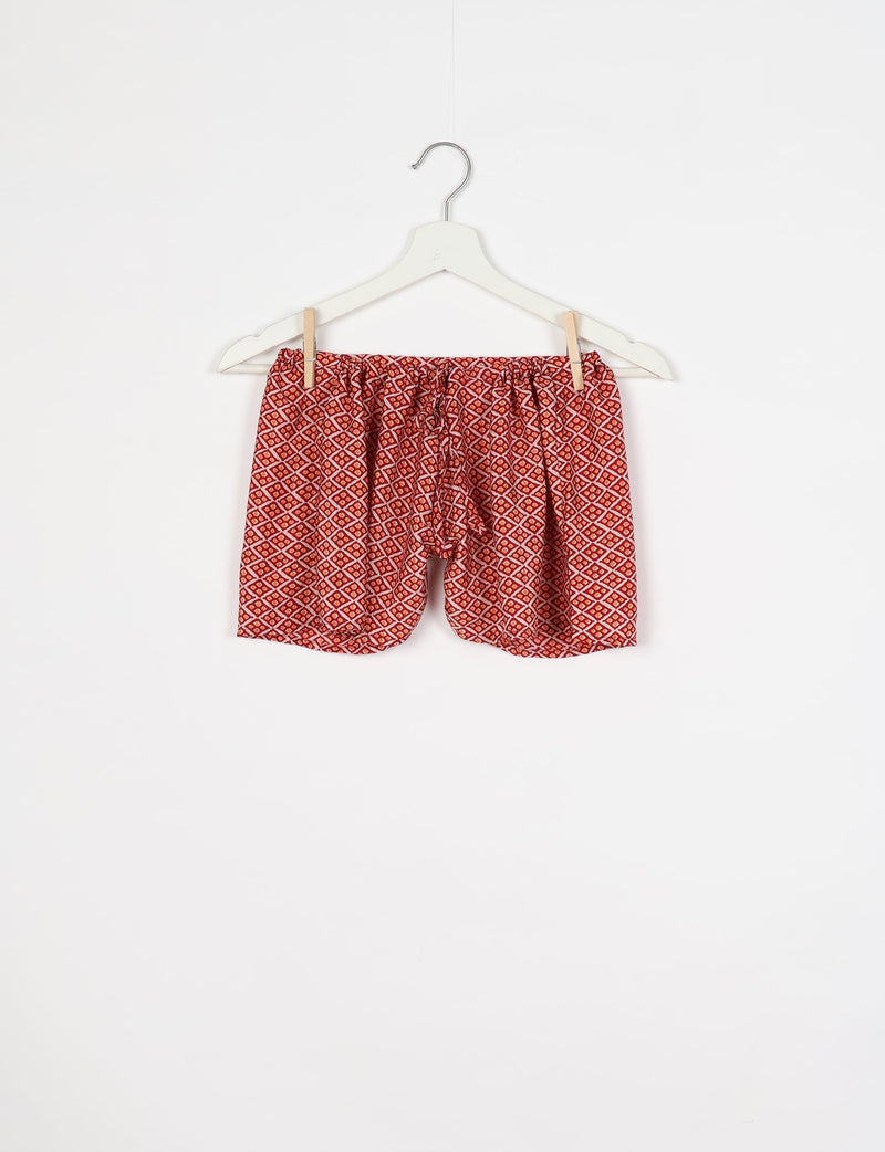 Red shorts with micro geometric diamond pattern hanging on white hanger with wooden clips against white background
