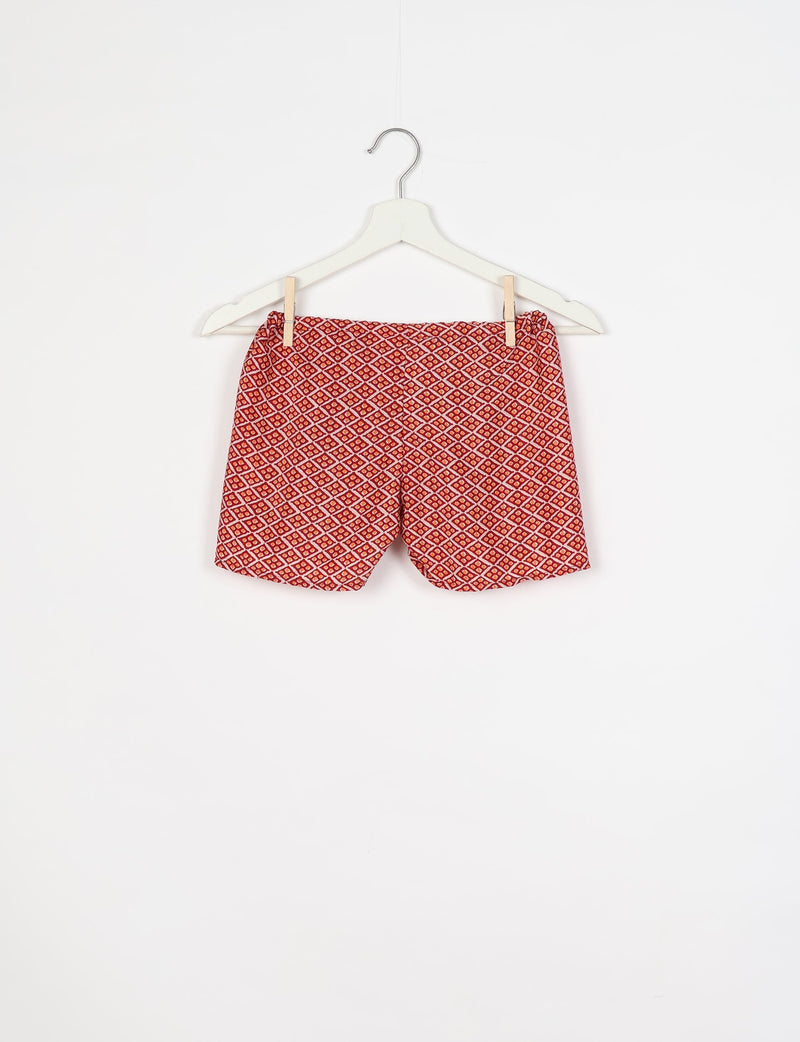 Red shorts with a micro diamond pattern in white and orange, hanging on a white hanger against a white background