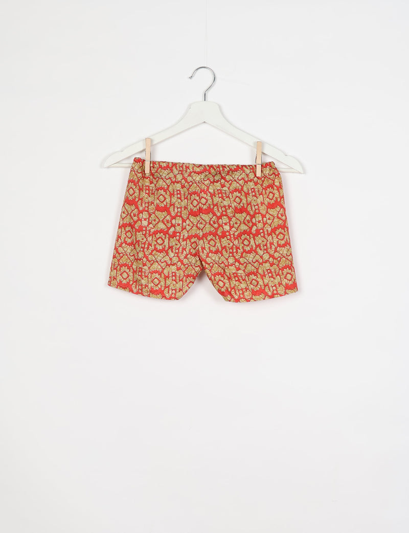 Orange shorts with beige and red ethnic pattern, elastic waistband, hanging on white hanger against white background