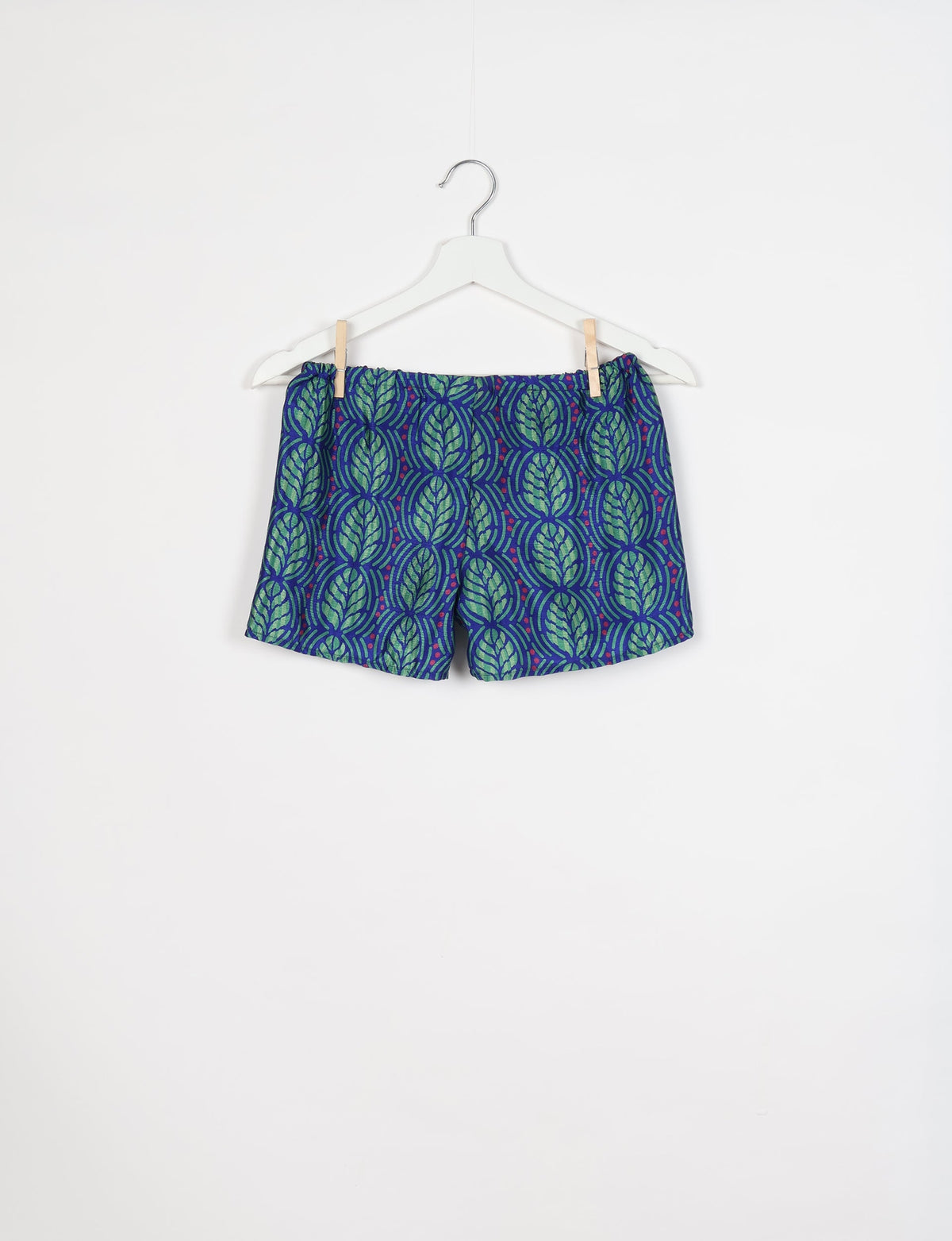 Blue shorts with green leaf pattern and small red dots, elastic waistband, hanging on white hanger against white background