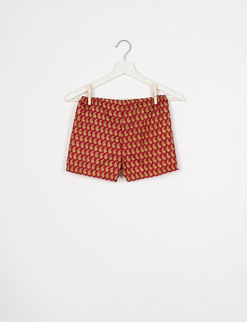 Red shorts with yellow paisley pattern hanging on white hanger against white background