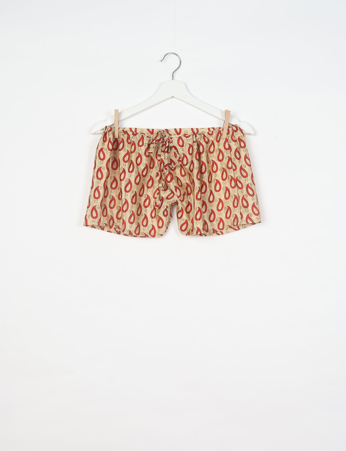 Beige pajama shorts with red paisley pattern and drawstring waist hanging on white hanger against white background