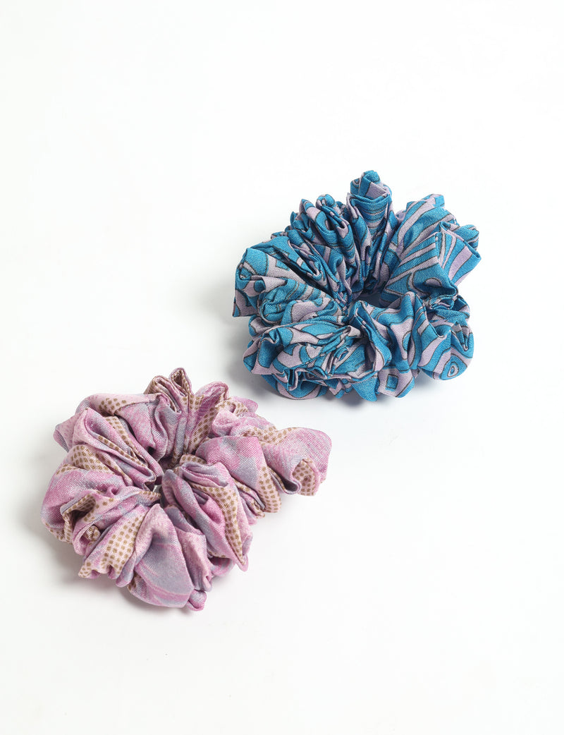 Set of two fabric scrunchies, one blue with purple and black abstract pattern, one pink with brown and purple pattern, flat lay