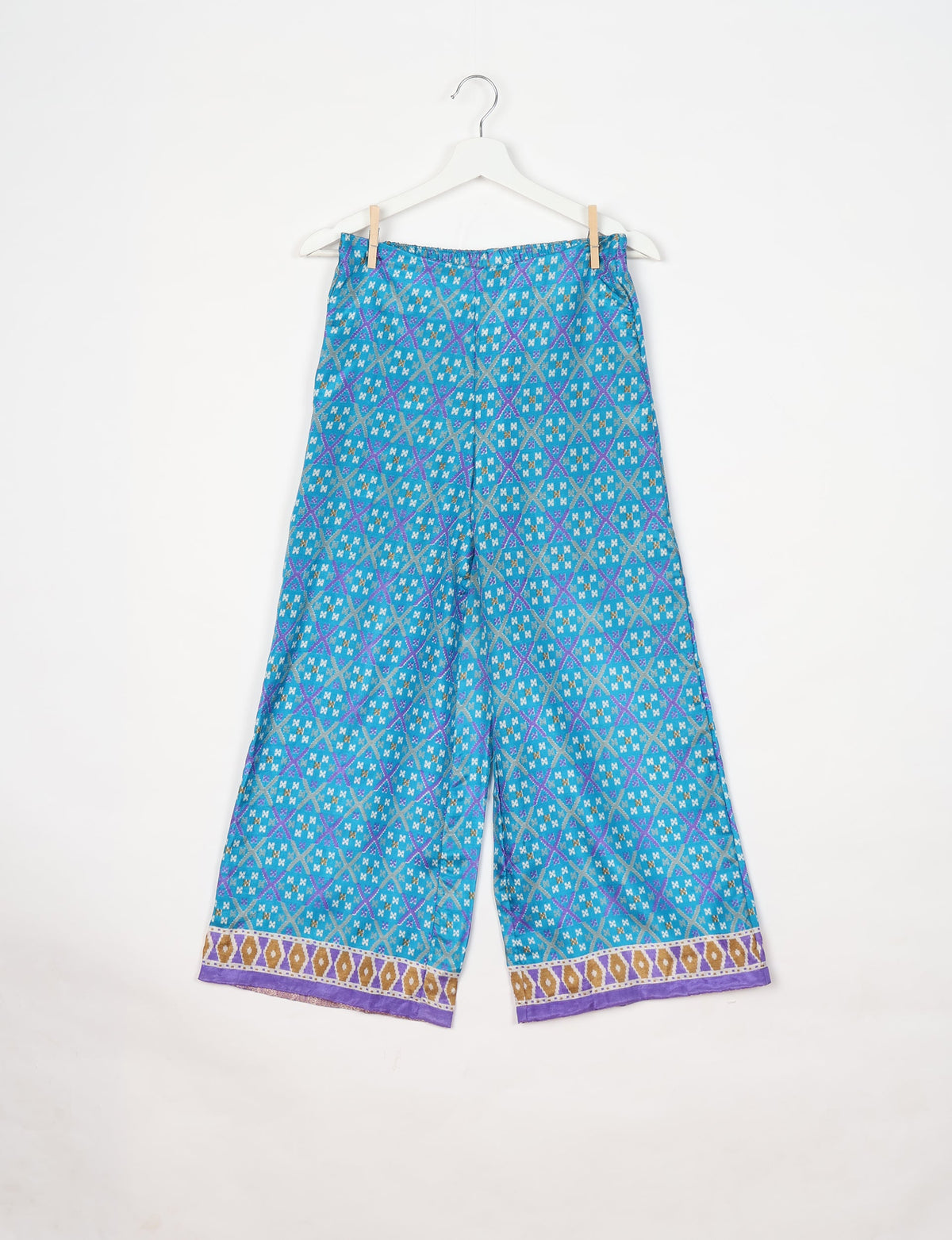 Blue geometric patterned wide-leg pants with elastic waistband and purple, gold, and white border detail, hanging on white hanger