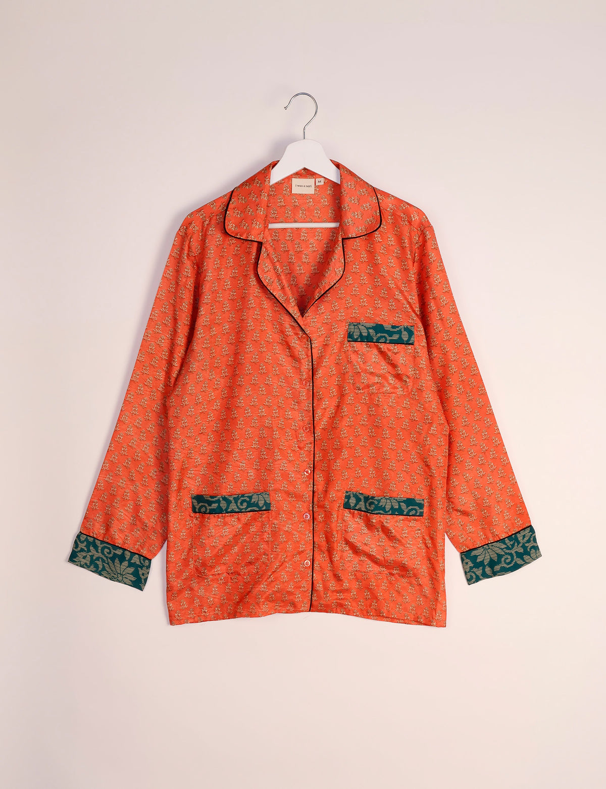 Orange long-sleeve pajama top with small floral print, dark green patterned cuffs and pockets, hanging on white hanger