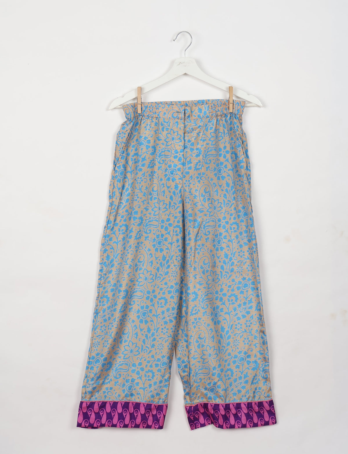 Blue and beige floral patterned pants with purple and pink patterned cuffs hanging on a white hanger against a white background