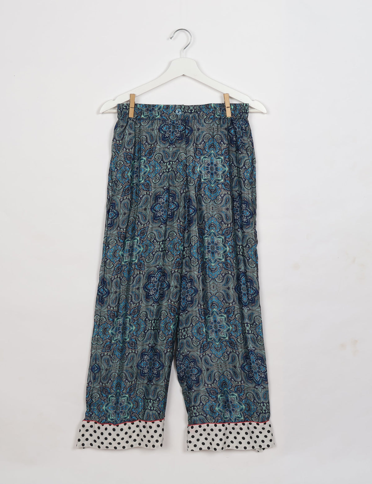 Blue and grey paisley patterned pajama pants with white polka dot cuffs hanging on a white hanger against a white background
