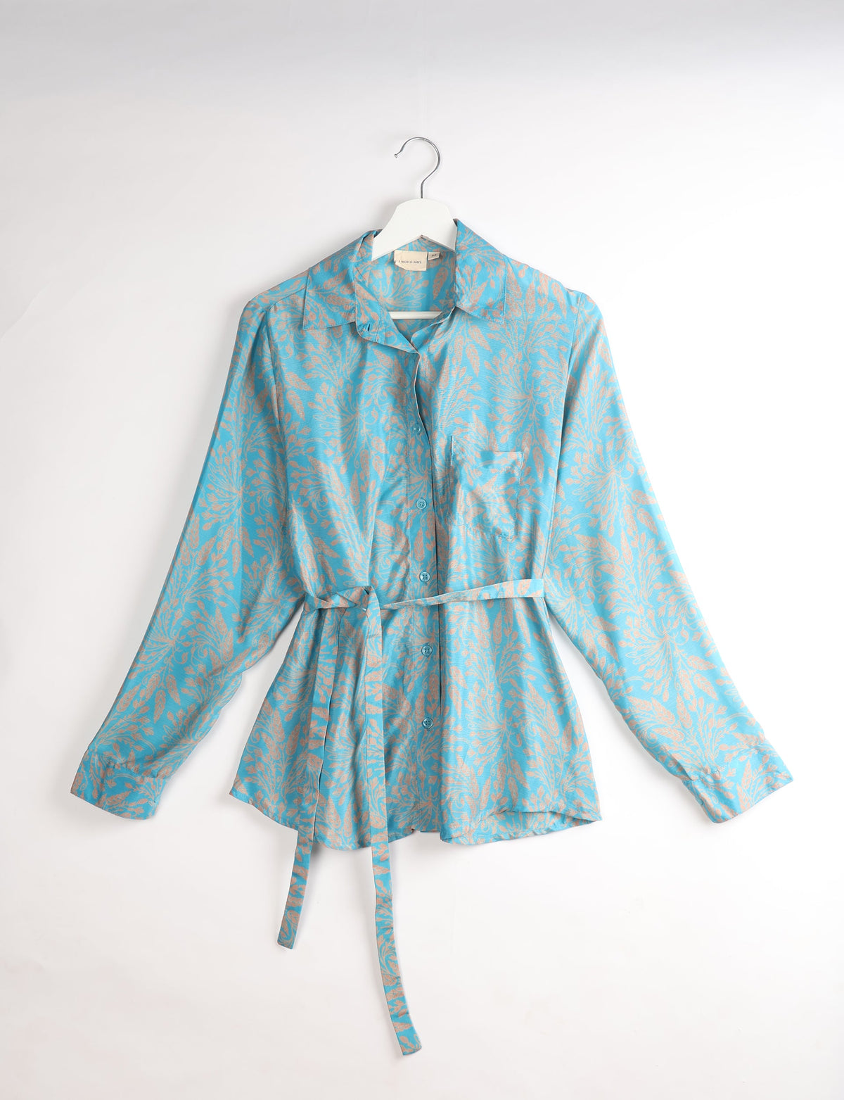 Blue button-up shirt with beige tropical leaf pattern, long sleeves, chest pocket, and matching fabric belt, hanging on