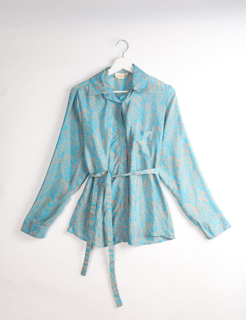 Blue button-up shirt with beige tropical leaf pattern, long sleeves, chest pocket, and matching fabric belt, hanging on