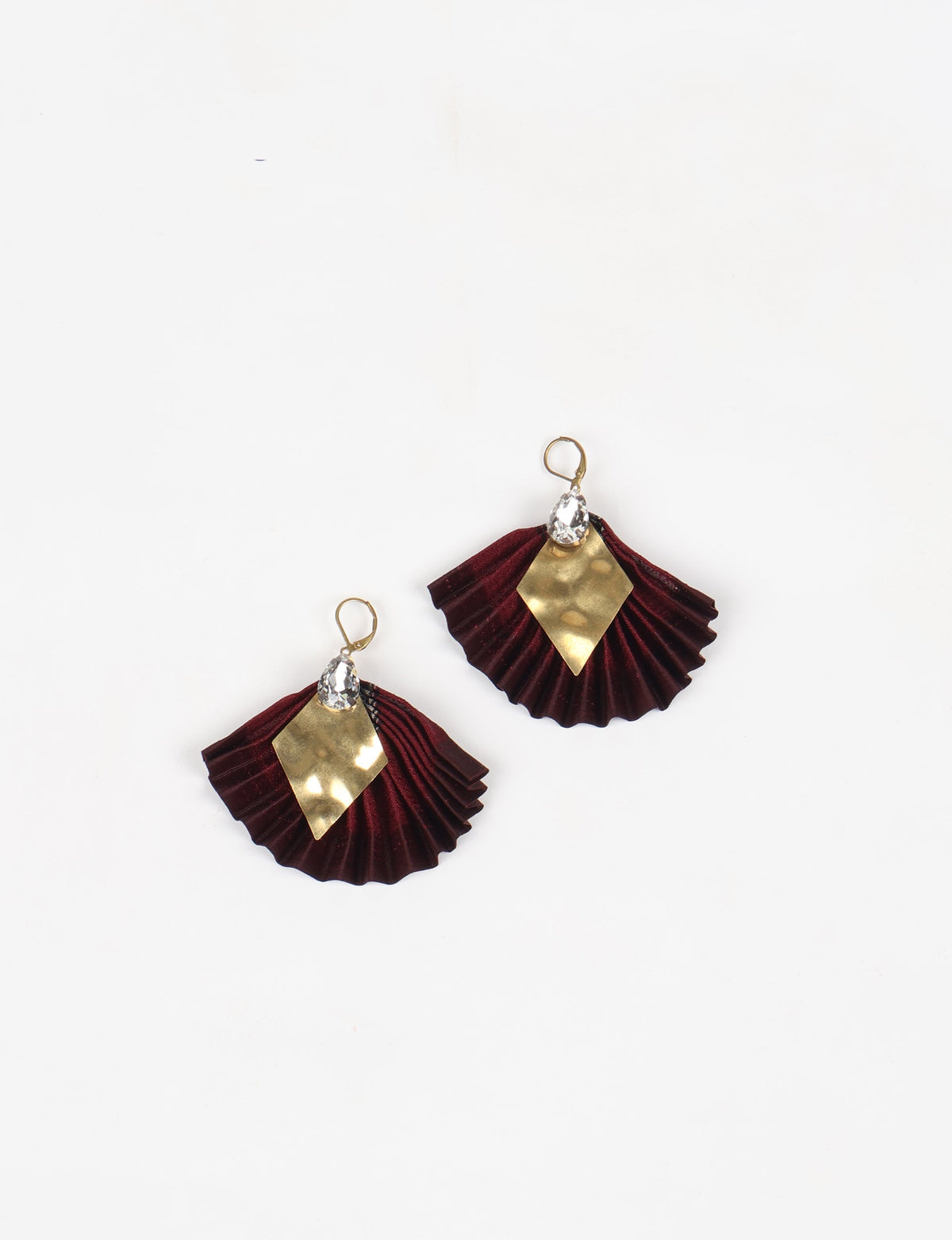 Pair of pleated earrings with deep red fabric, gold diamond-shaped metal accents, and clear teardrop crystals, flat lay view