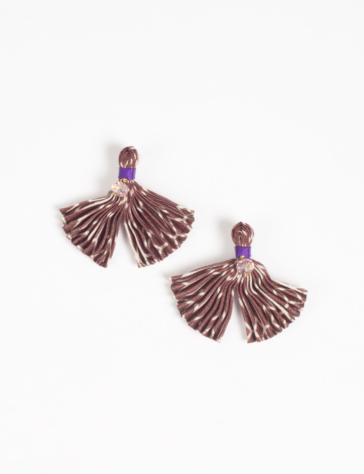Beige and brown pleated fabric earrings with abstract pattern, purple band, and small clear bead, flat lay on white background