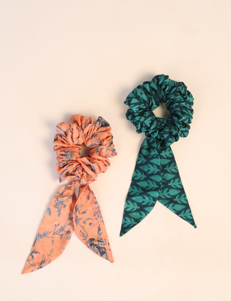 Set of two scrunchies with attached tails, one orange with gray floral print and one green with dark leaf pattern, flat lay