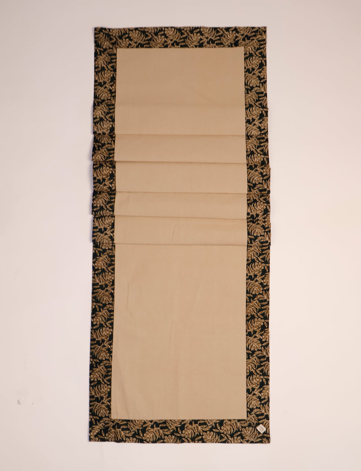 Beige table runner with black border featuring a gold leaf pattern, shown flat on a light background