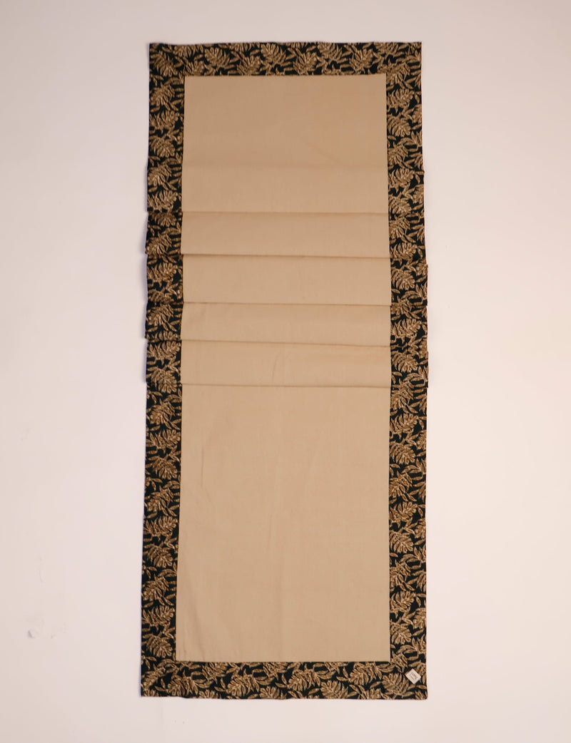 Beige table runner with black border featuring a gold leaf pattern, shown flat on a light background