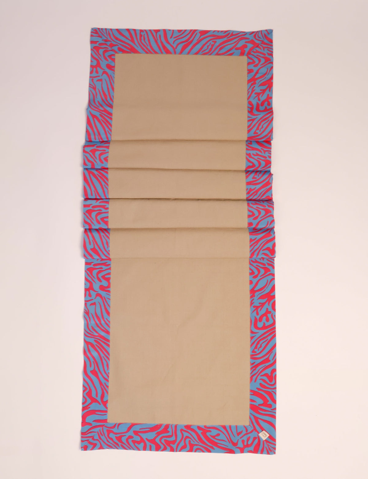 Beige table runner with blue and red abstract patterned border, shown flat with a small fabric tag on corner