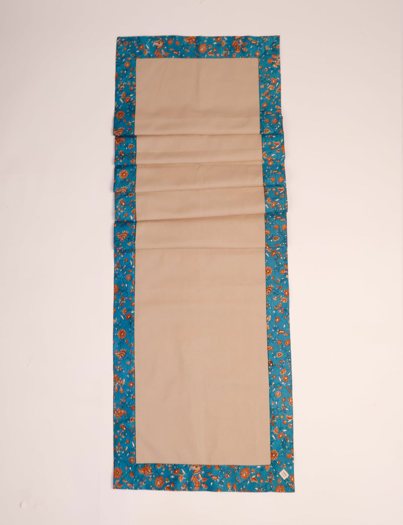 Beige table runner with blue tropical floral border, flat laid on a light background, small label on corner visible