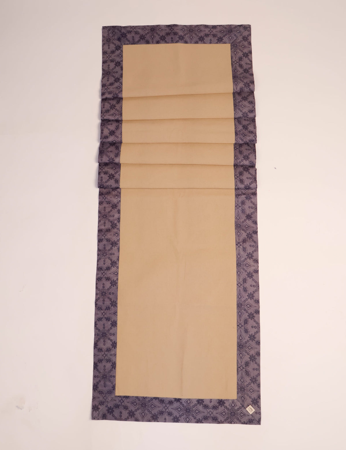 Beige table runner with purple ethnic patterned border, flat laid with visible fabric texture and a small label on corner