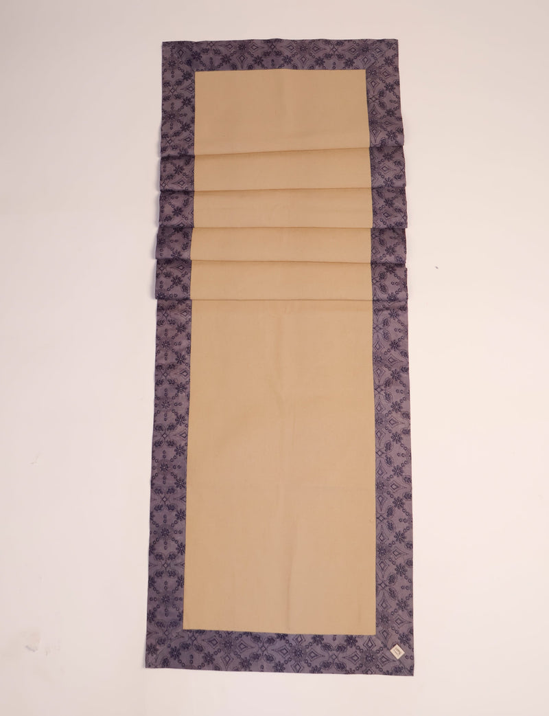 Beige table runner with purple ethnic patterned border, flat laid with visible fabric texture and a small label on corner