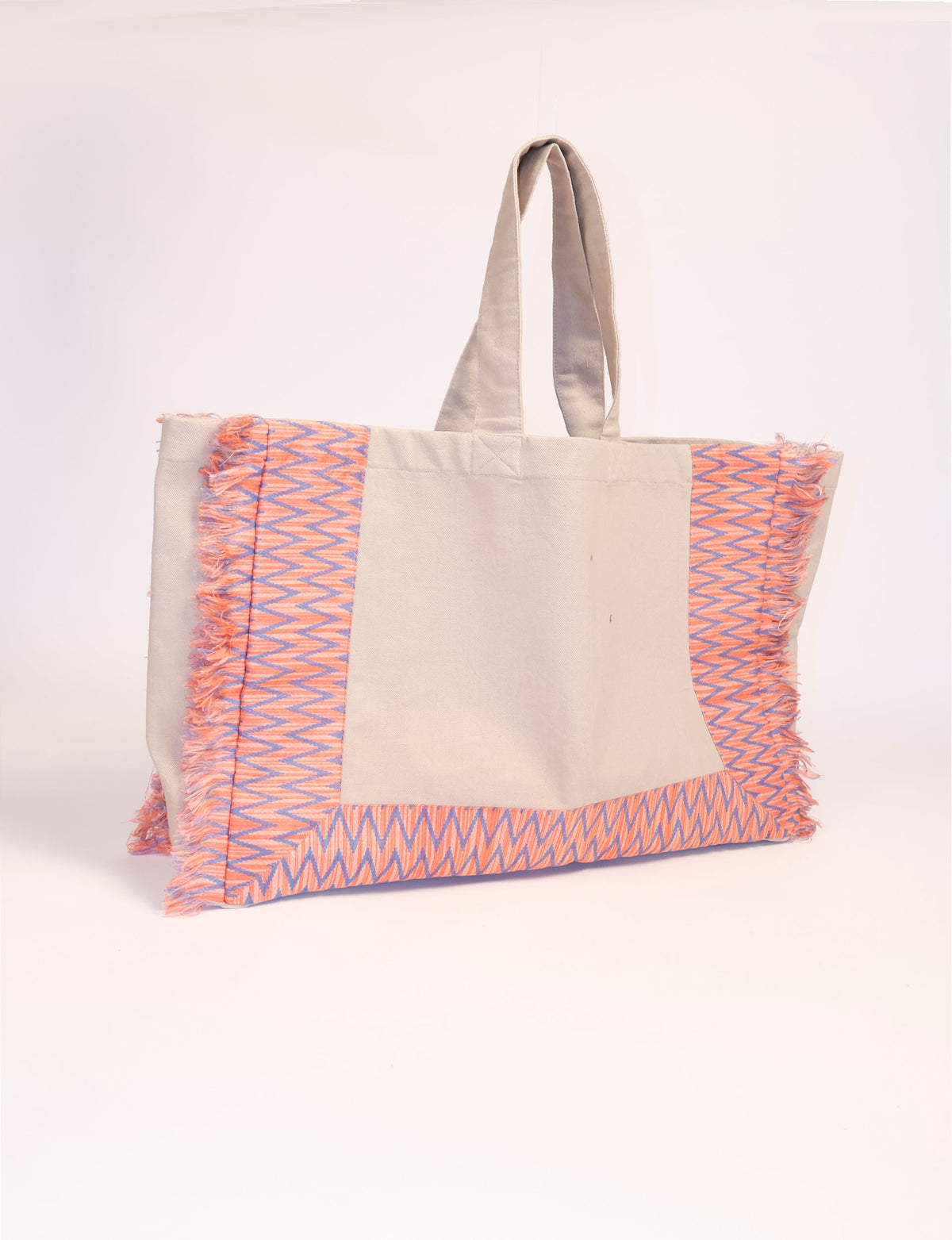 Beige fabric carry-all bag with orange and purple zigzag geometric fringe trim, shown hanging by handles