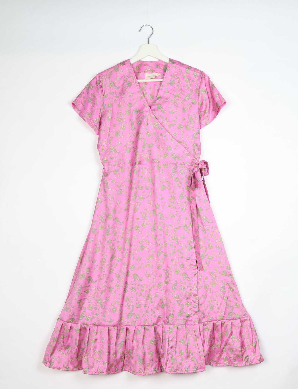 Pink midi wrap dress with tropical floral and paisley print, short sleeves, side tie, and ruffled hem, hanging on white