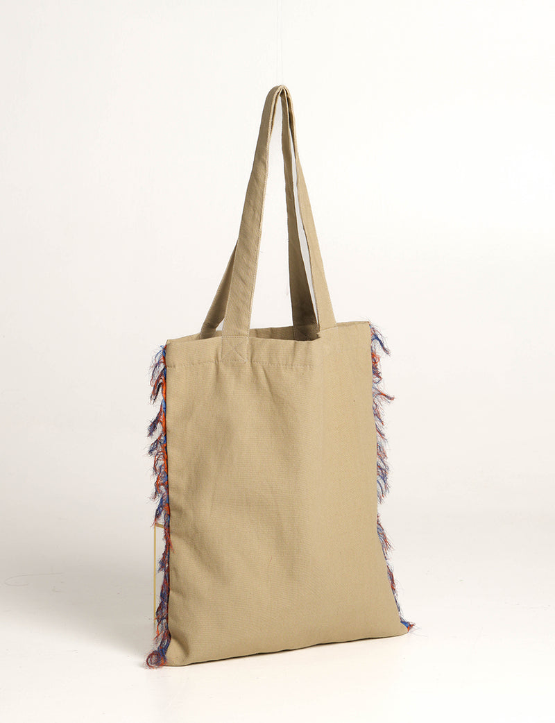 Beige fabric shopper bag with long handles and blue and orange fringe details on the sides, hanging flat