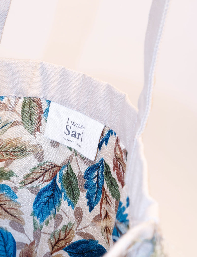 Beige shopper bag with blue and brown leaf pattern fabric interior and white woven handles, label reading "I was a Sari" visible