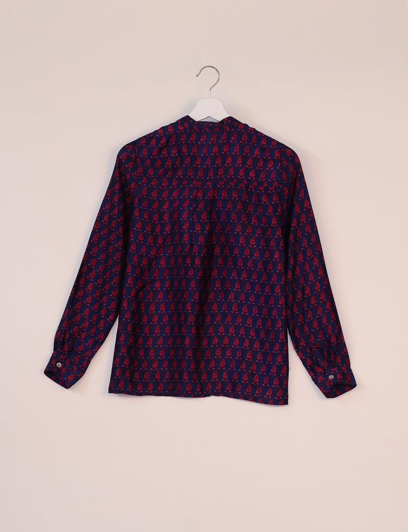 Blue blouse with red paisley micro design, long sleeves, buttoned cuffs, shown from back on white hanger against beige wall