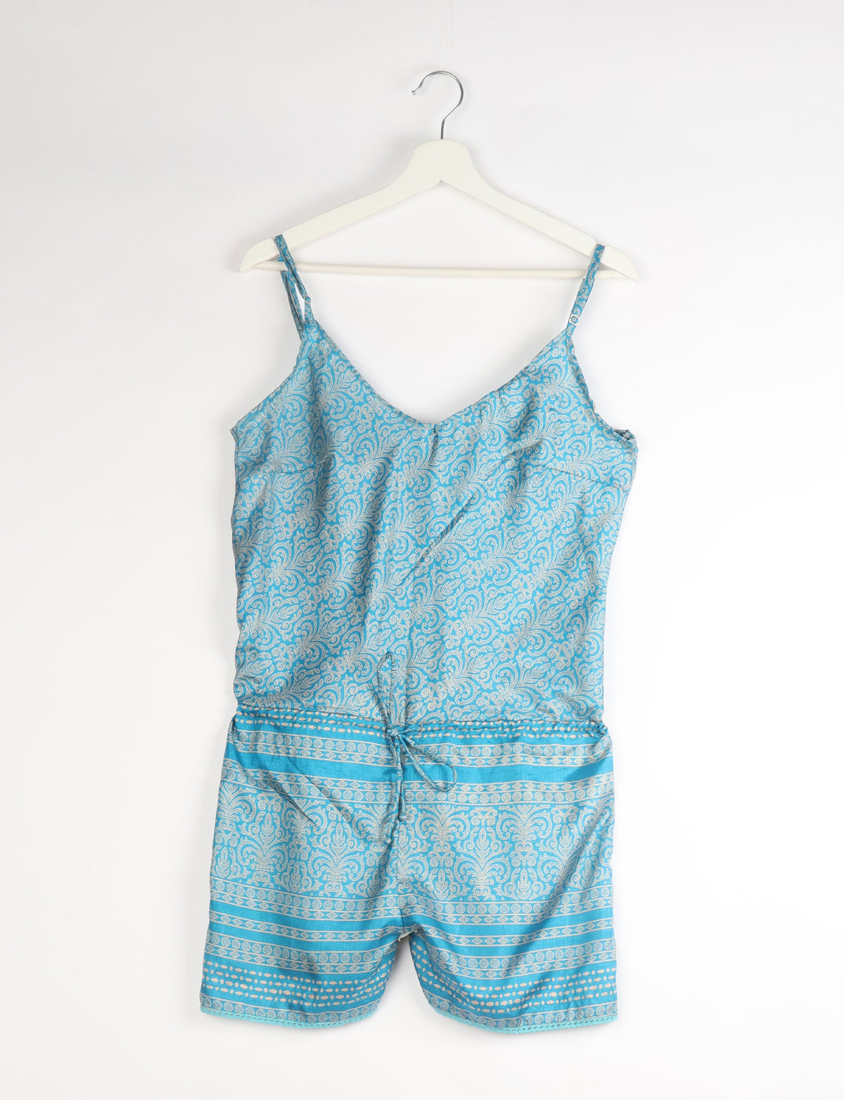 Blue playsuit with white abstract paisley and stripe patterns, adjustable spaghetti straps, and a front tie, hanging on